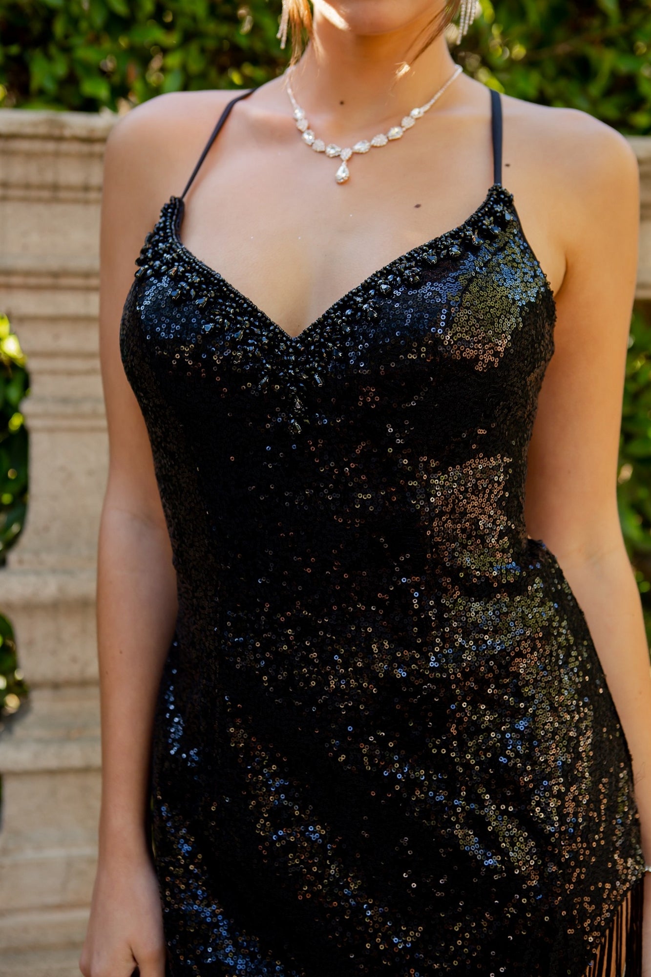 A woman wearing the Long Sequin Formal Prom Dress with Fringe 8036J and a pearl necklace stands outdoors in front of stone and greenery.